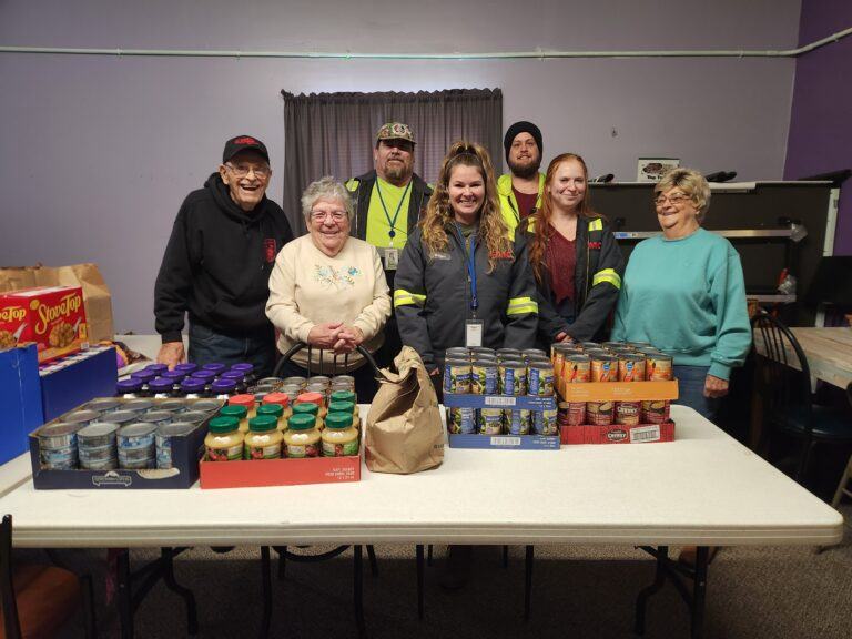 Volunteers stand behind a table of donated canned goods, snacks, and pantry items collected during a middleport food drive.